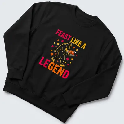 Bigfoot carrying a turkey with the text 'Feast Like A Legend' in colorful cartoon style. — printed on a unisex sweatshirt